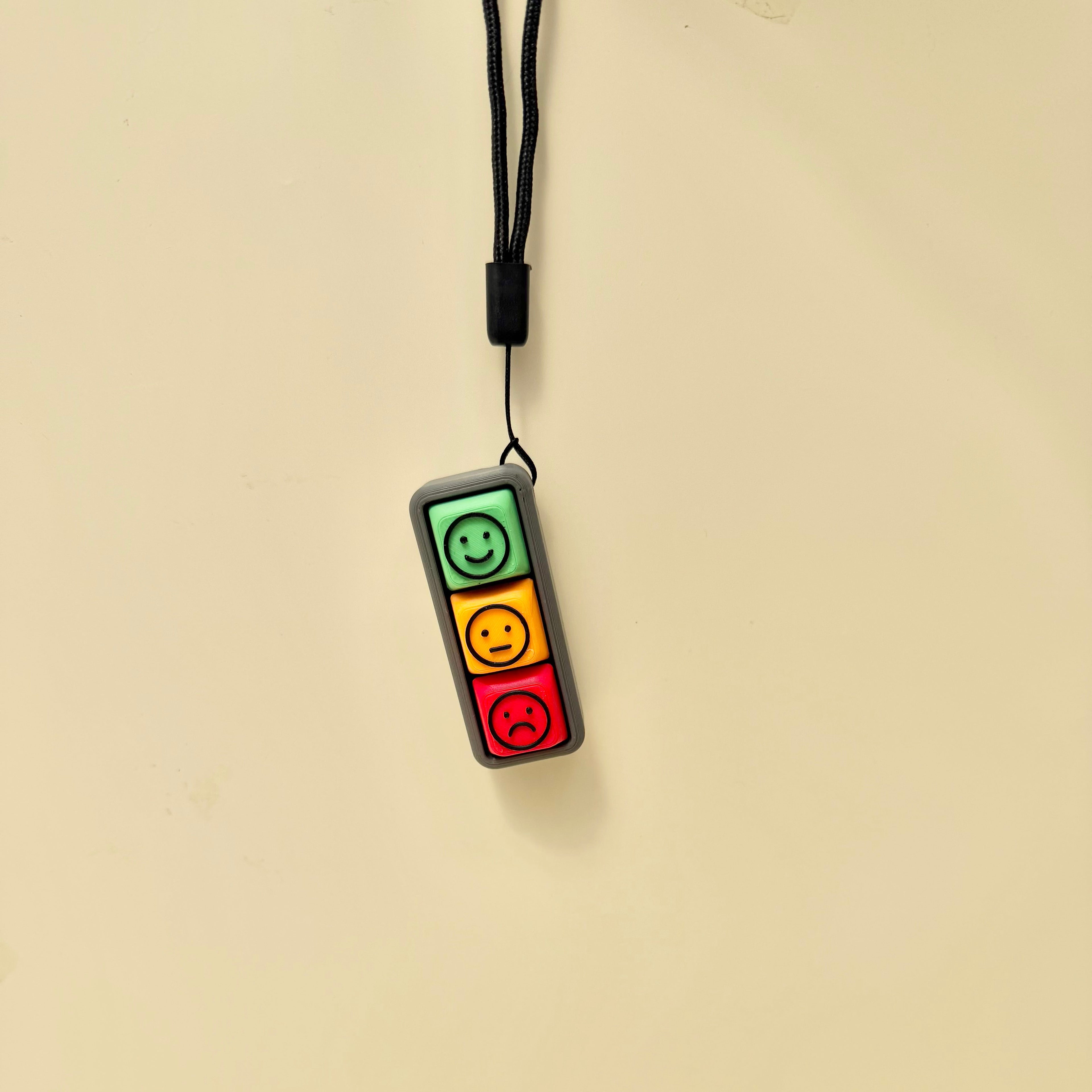 Three-button mood fidget with visual face symbols, tactile click feedback and wrist strap attachment.