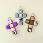 Three colorful cross-shaped fidget keychains with various designs on a beige background