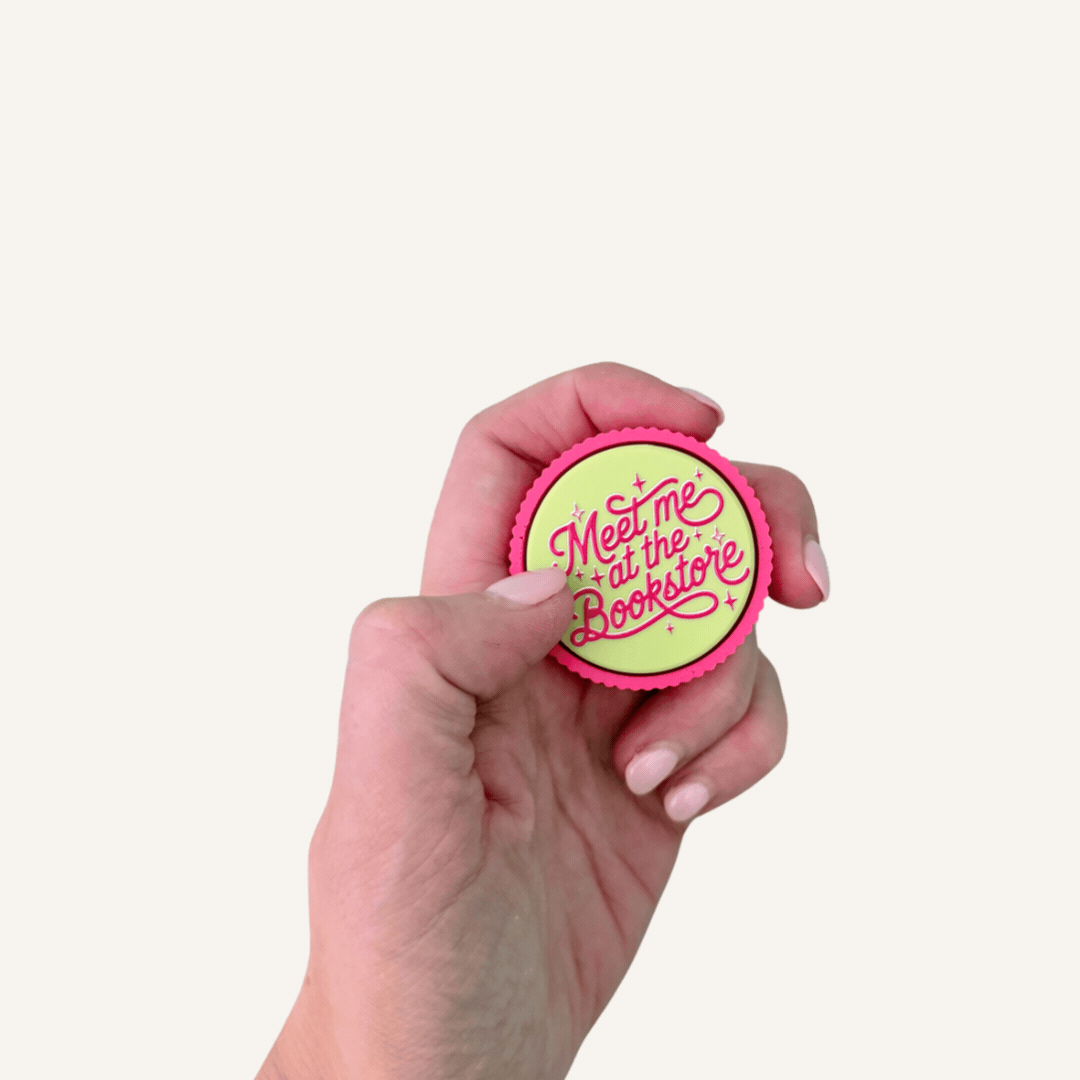 Pink and lime Meet Me at the Bookstore sensory clicker with raised lettering and tactile ridged edge.